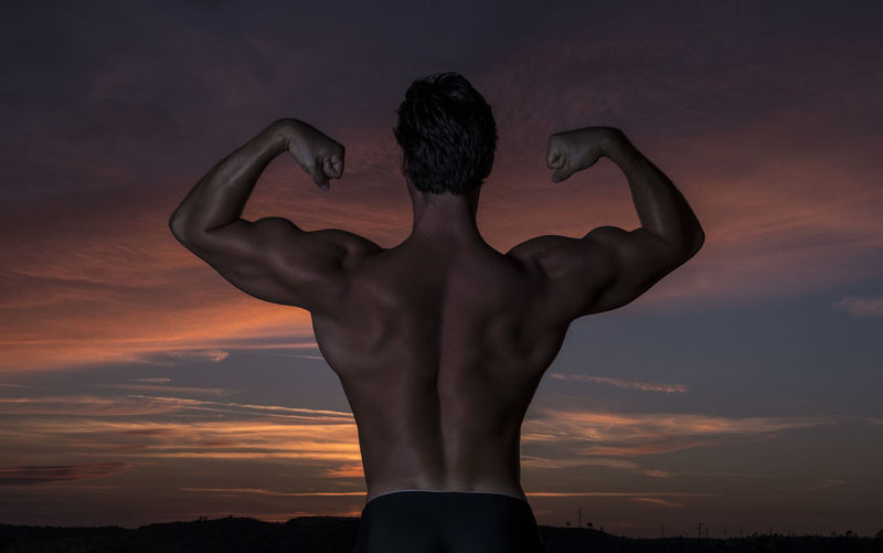 Rear view of shirtless man flexing muscles against sky during sunset