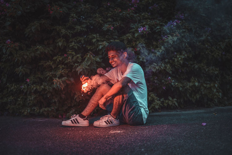 Young man looking away while sitting on plants at night