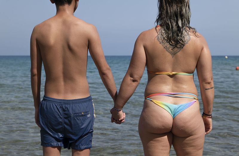 Rear view of girl in bikini and teenager boy in swimwear holding hand together standing on beach