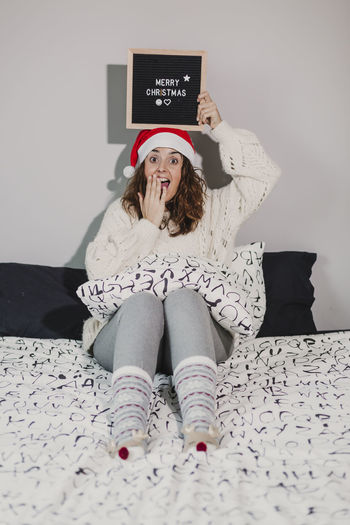 Portrait of woman with merry christmas text sitting on bed at home
