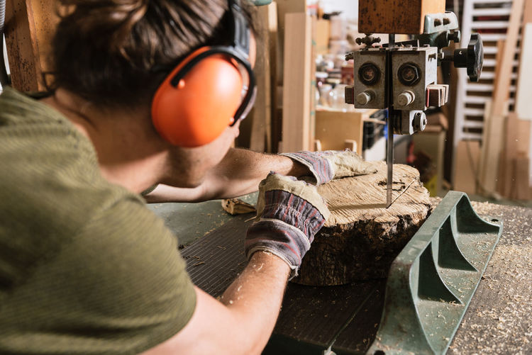 Male carpenter in protective ear muffs and goggles cutting piece of wood with band saw in shabby workshop
