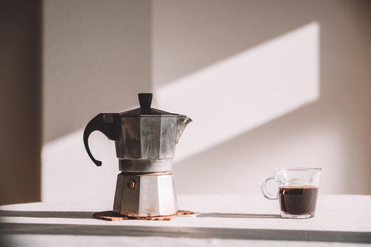 Metal geyser coffee maker placed on table with aromatic coffee served in irish glass in kitchen against white wall on sunny day