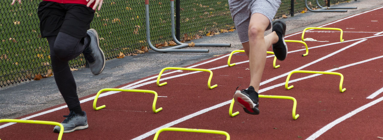 Two boys running over yellow mini hurdles on a red track during track and field practice outdoors.