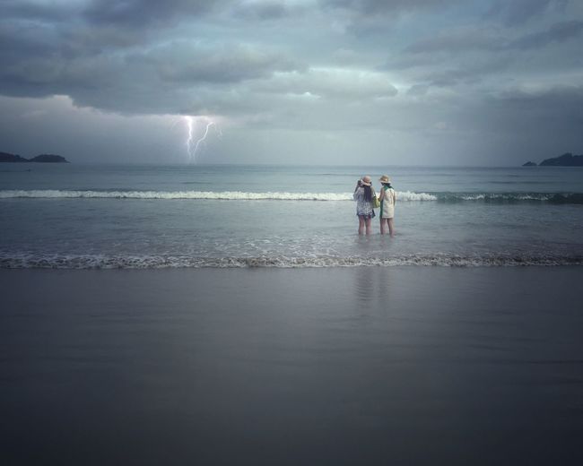 Rear view of women standing at beach against storm clouds