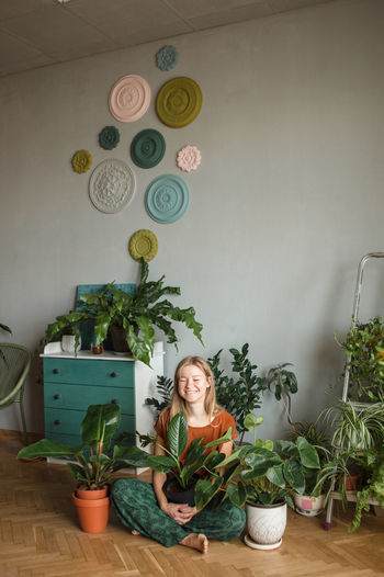 Happy woman sits in lotus pose on floor surrounded by potted plants