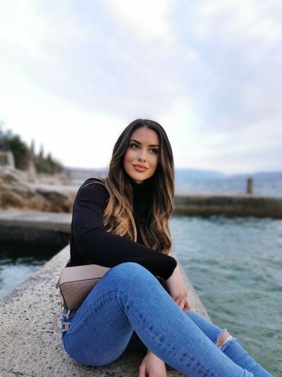 Beautiful young woman sitting by sea against sky