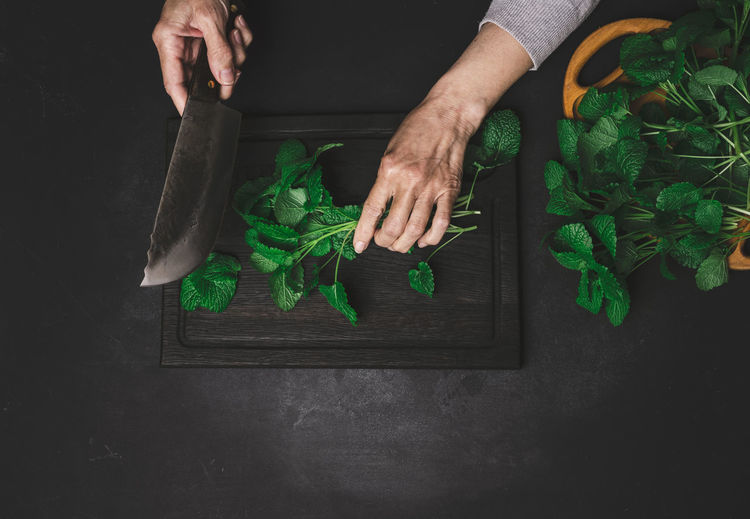 Woman cutting green mint leaves on brown wooden cutting board, top view