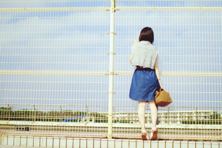 Rear view of girl looking through metal fence against sky