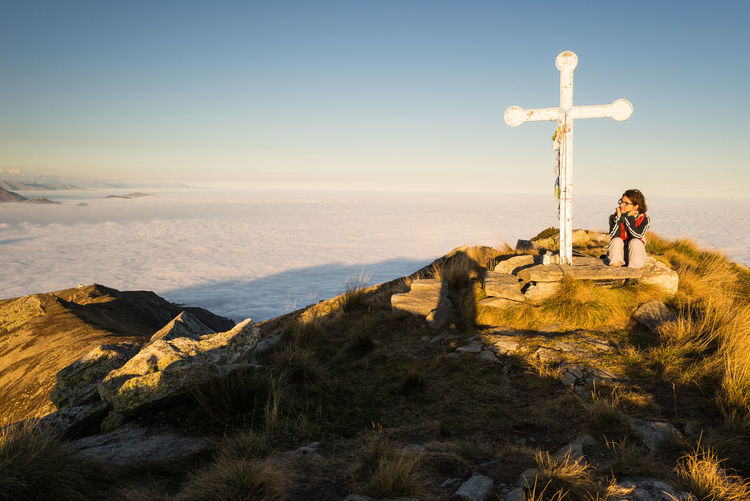 Female hiker sitting by cross against sky on mountain