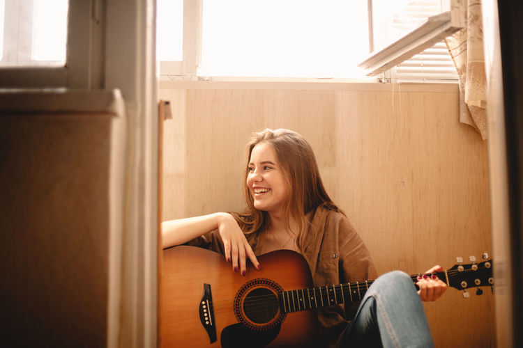 Cheerful young woman holding guitar while sitting on balcony floor