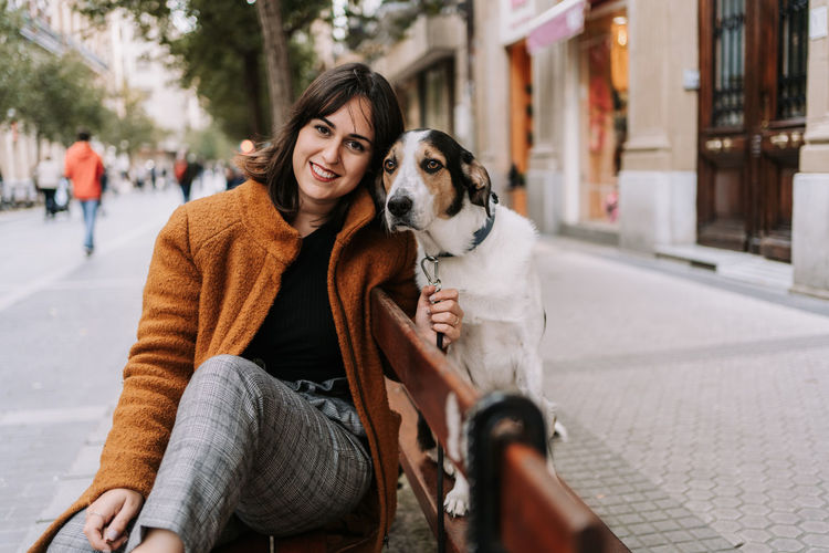 Cheerful female sitting on wooden bench with friendly treeing walker coonhound dog on leash and looking at camera