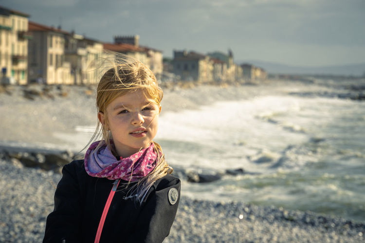 Girl posing in front waterfront against sky, breaking waves on rocky beach, marina de pisa