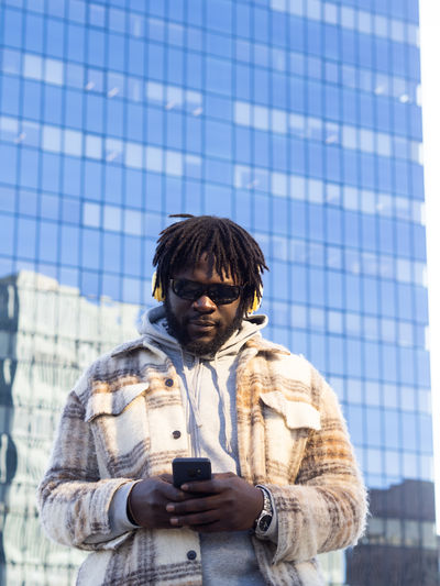 Trendy african american male in sunglasses with headphones and cellphone listening to music while standing with hand in pocket against building reflecting town