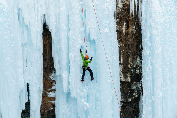 Male ice climber dangles from frozen waterfall climbing up the challenging landscape in victory. 