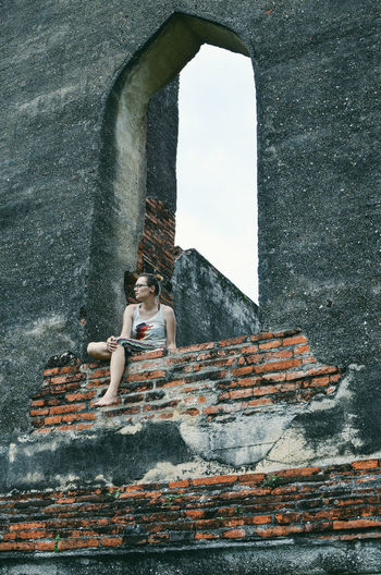 Low angle view of woman sitting on old brick wall