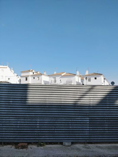 Low angle view of metal wall and buildings against clear blue sky