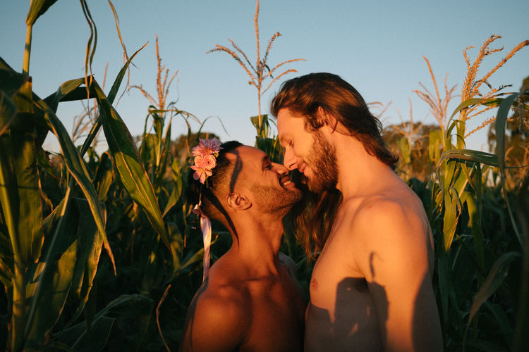 Side view of shirtless gay couple romancing while standing amidst plants