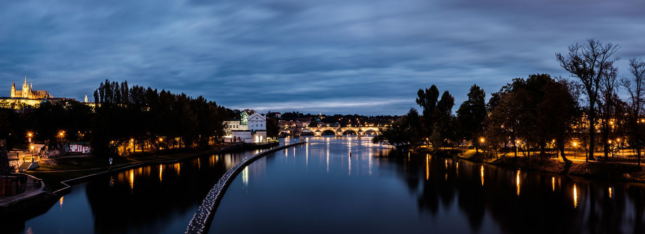 Panoramic view of river by illuminated city against sky at dusk