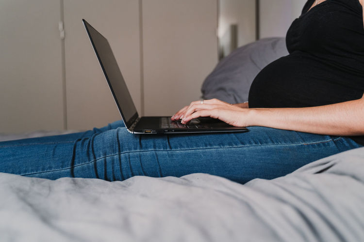 Midsection of pregnant woman using laptop while sitting on bed at home