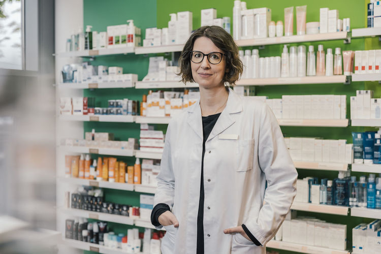 Confident pharmacist with hands in pockets standing in chemist shop