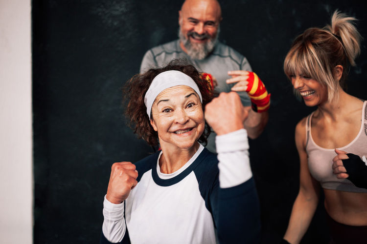 Optimistic senior woman in sportswear standing in boxing position and looking at camera while having fun with friends during training in gym