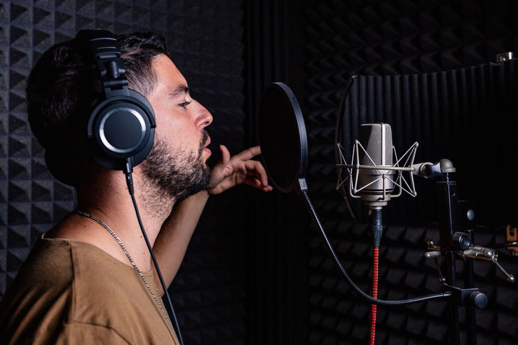 Side view of male singer in headphones standing in acoustic room with soundproof walls and microphone and recording song in studio