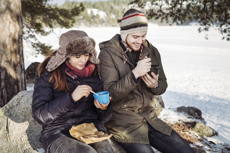 Couple having food while sitting on rock at field during winter