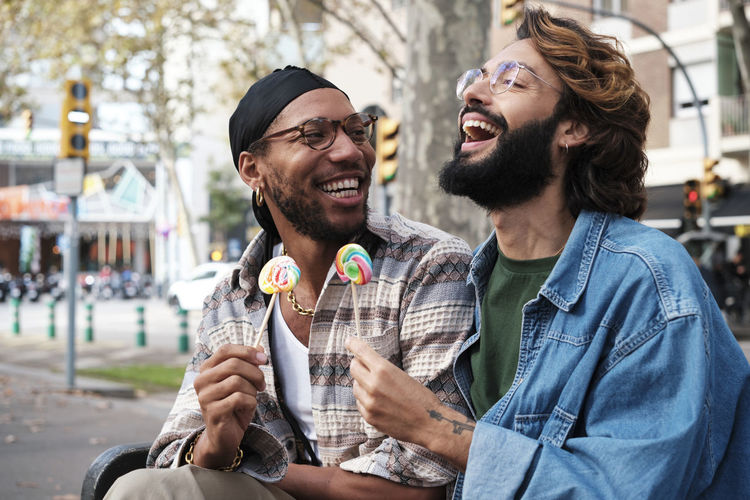 Happy gay couple enjoying candy sitting on bench