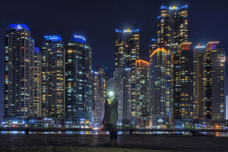 Woman holding flash light while standing against illuminated buildings in city at night