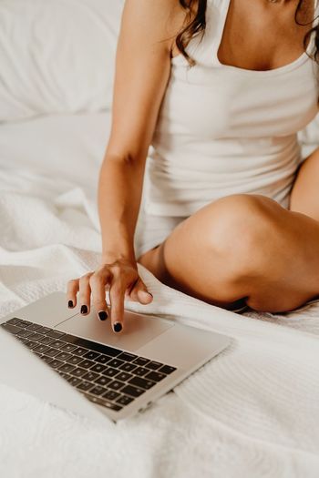 Cropped unrecognizable delighted young female in sleepwear sitting on bed and browsing laptop after awakening in light bedroom in morning