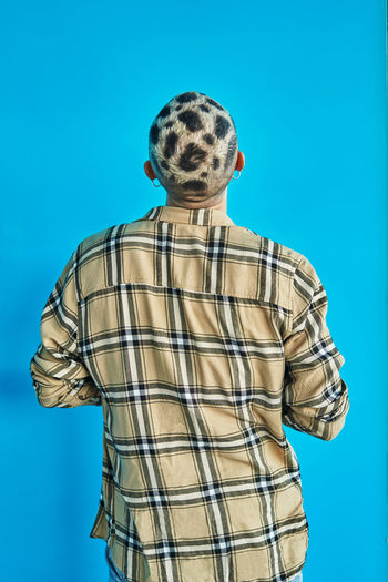Back view of anonymous male with trendy eccentric haircut wearing checkered shirt standing against blue background