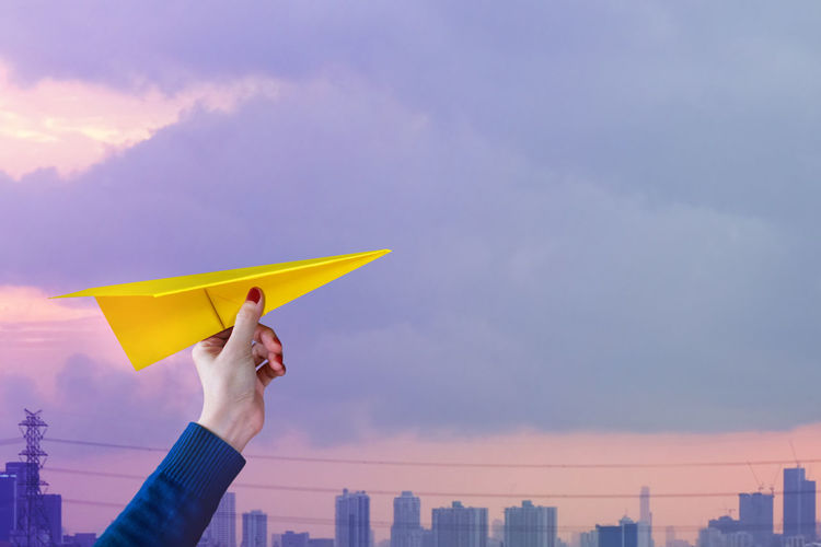 Cropped hand of woman holding paper plane against cloudy sky during sunset