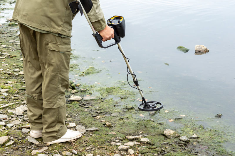 A man with a metal detector on the shore of a reservoir.