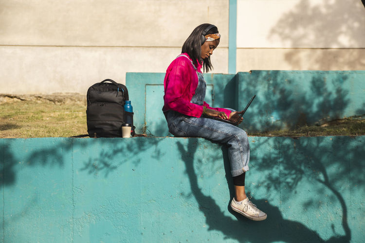Young woman using laptop by backpack on wall