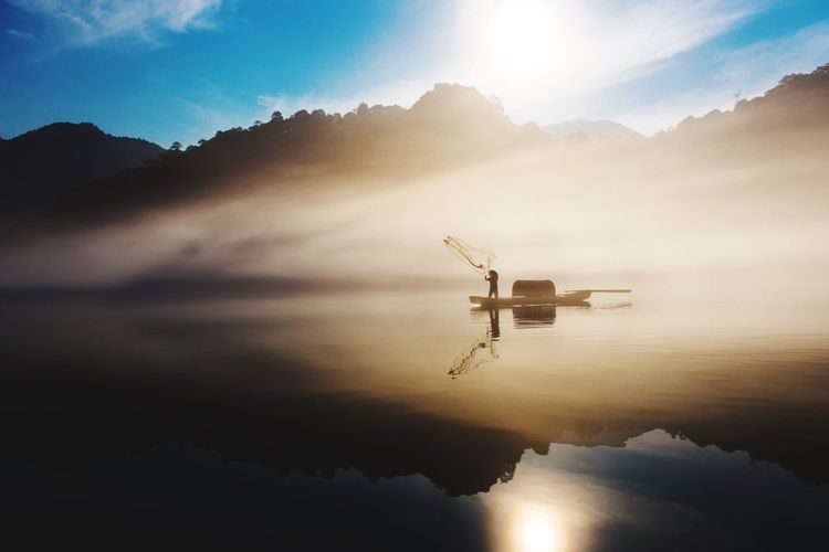 Silhouette fisherman throwing fishing net on lake in foggy weather at sunrise