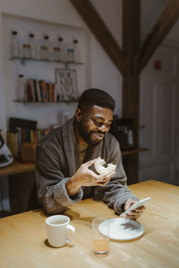 Smiling man using smart phone and having doughnut at home