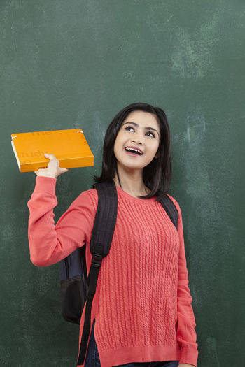 Happy female student holding book while standing by blackboard