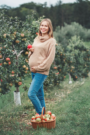 Portrait of smiling pregnant woman holding apple while standing by basket and trees