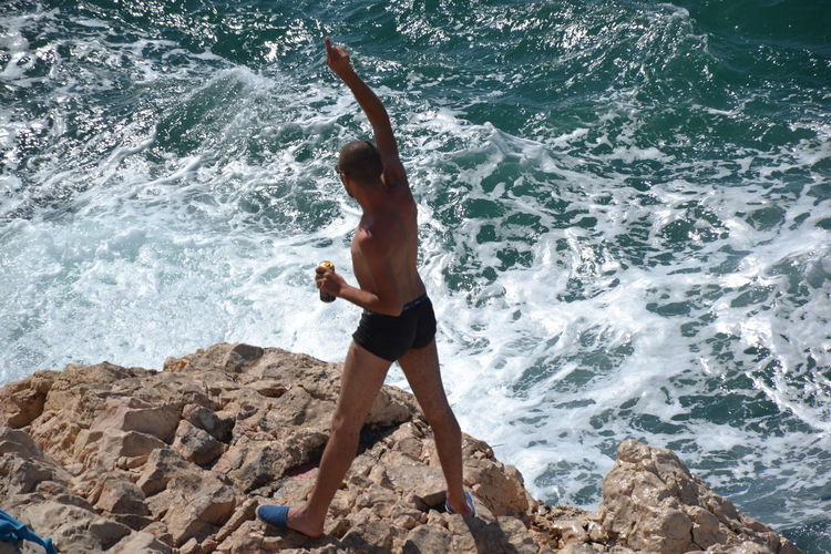 High angle view of shirtless man pointing while standing on rocky shore