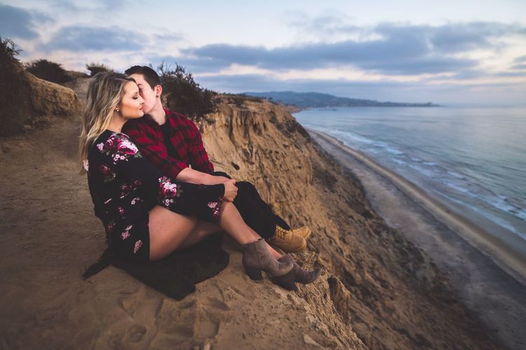 Romantic couple sitting on cliff by sea against cloudy sky