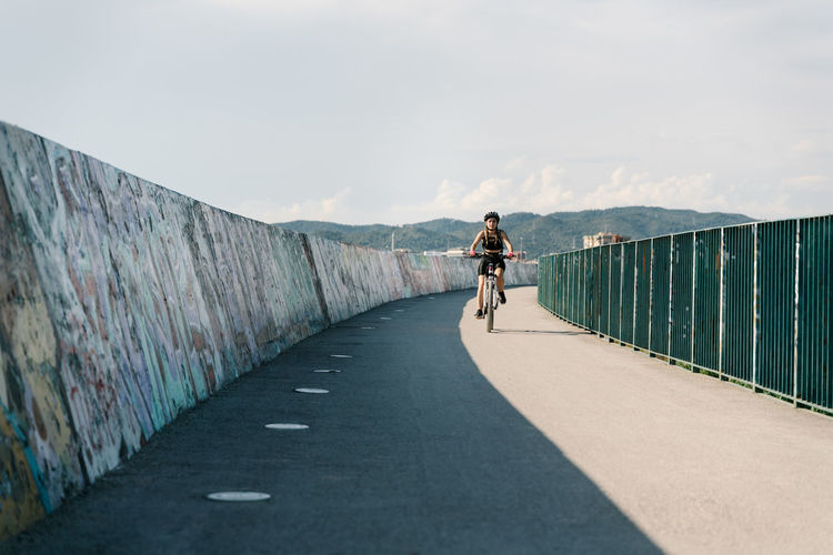 Low angle of confident young female bicyclist in sportswear and helmet riding bike on fenced curvy paved track while training alone in summer day
