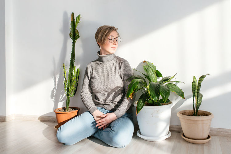Young woman sitting on potted plant against wall at home