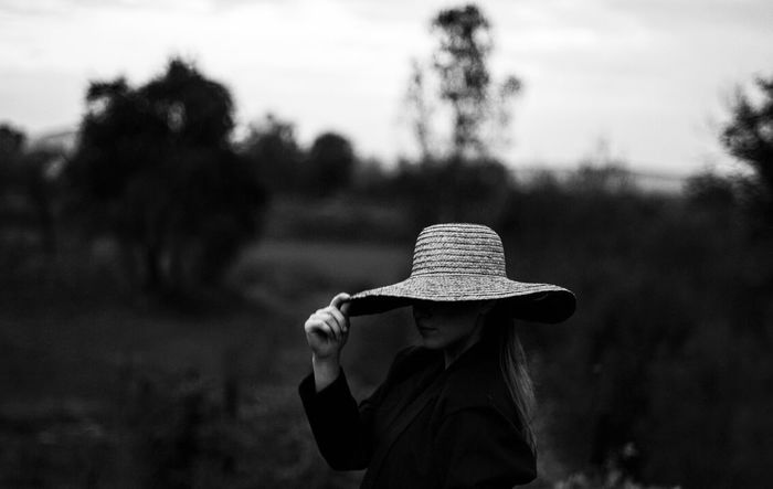 Woman holding hat while standing on field against sky