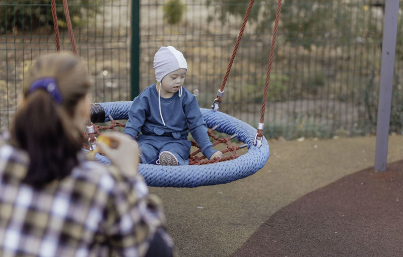 Cute little boy with down syndrome in a funny hat with his mother, swinging on a swing, soap bubbles