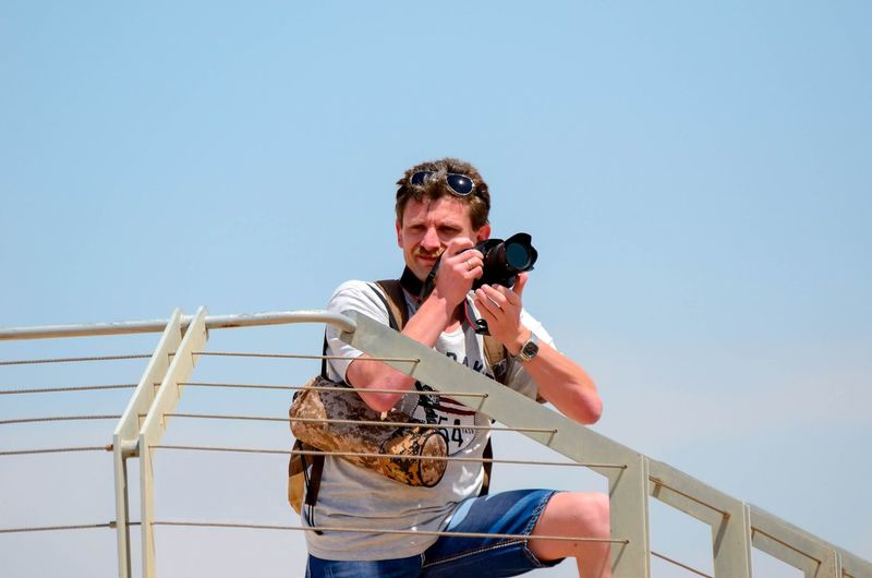 Low angle view of man looking away while photographing by railing against sky