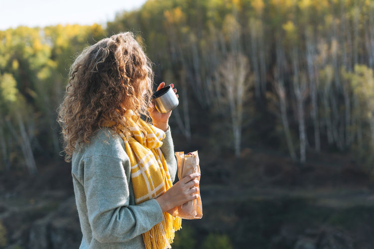 Young happy beautiful woman traveller with curly hair eating hot dog and drinking tea on nature view