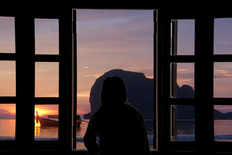 Rear view of silhouette woman looking through window at sunset