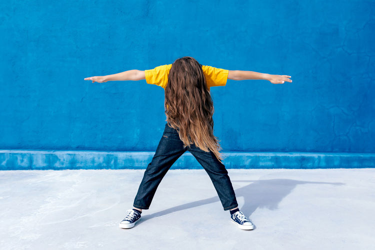 Anonymous energetic teenager in cool clothes dancing with outstretched arms on blue background