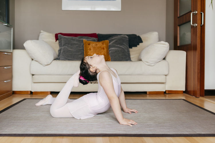 Side view of little girl in leotard looking up touching head with feet while doing gymnastic exercise on floor in cozy living room