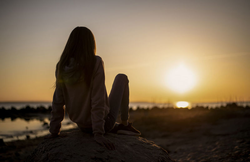 Rear view of girl sitting at beach during sunset
